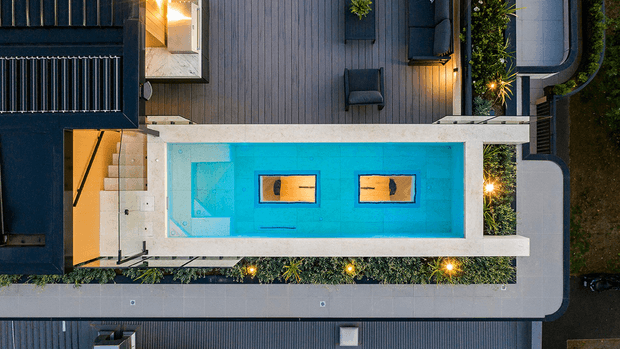 Aerial top-down view of a contemporary residential outdoor area centred on a long rectangular swimming pool with bright turquoise water and two in-water loungers or platforms. The pool is bordered by pale concrete coping, timber decking, integrated steps, dark lounge furniture, planted garden beds, architectural walls, and curved paved walkways. Warm landscape lighting creates a refined evening atmosphere.
