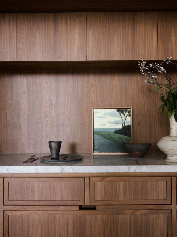 Close-up interior vignette of a refined timber built-in dining or serving cabinet. The wall and cabinetry are clad in richly grained medium-to-dark timber panels, with a pale veined stone countertop, integrated drawers and framed cupboard fronts. A dark ceramic place setting, framed landscape artwork, bowl and large vase provide restrained styling. The composition is warm, polished and highly detailed, with no wider room context visible.