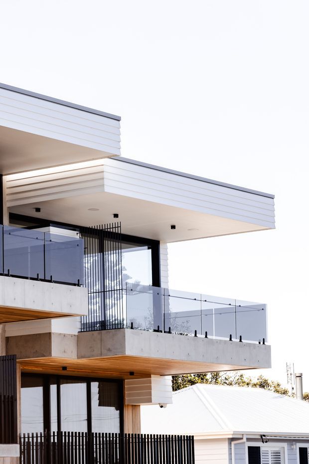 Exterior architectural photograph of a contemporary multi-level residence featuring crisp horizontal white cladding, broad flat roof overhangs, concrete balconies, clear glass balustrades, black metal screening and fencing, and warm timber soffit lining. A smaller pitched-roof building is visible in the background.
