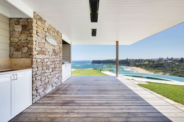 A covered outdoor entertaining deck overlooking a bright coastal bay and sandy beach. The space features a built-in outdoor kitchen with white cabinetry and a stone-clad wall, timber decking, a slim timber post, frameless glass balustrading, and a white slatted ceiling with recessed linear lighting. A swimming pool and landscaped lawn are visible beyond the deck.