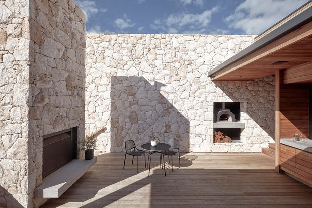 Sunlit contemporary outdoor courtyard with pale irregular stone walls, a built-in fireplace, timber decking, and a partially covered outdoor kitchen or entertaining area. The courtyard includes a small black café-style table and chairs, with strong architectural shadows and a warm timber soffit creating a refined, relaxed setting.