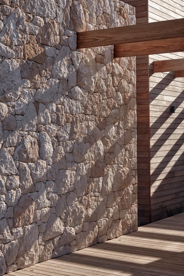Close-up architectural detail of a rustic stone masonry wall meeting a timber-clad structure and timber deck. Heavy exposed wood beams project across the stonework, creating a warm, tactile composition with strong sunlight and shadows. No interior room is visible; the image focuses on exterior materials and construction details.