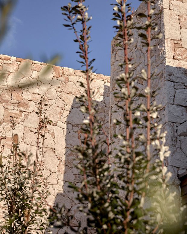 Exterior architectural detail of a sunlit courtyard or garden enclosure formed by rough pale natural-stone masonry walls, with narrow vertical wall sections and a partial opening at right. Tall leafy planting is prominently blurred in the foreground against a clear blue sky, creating a warm, rustic Mediterranean atmosphere.