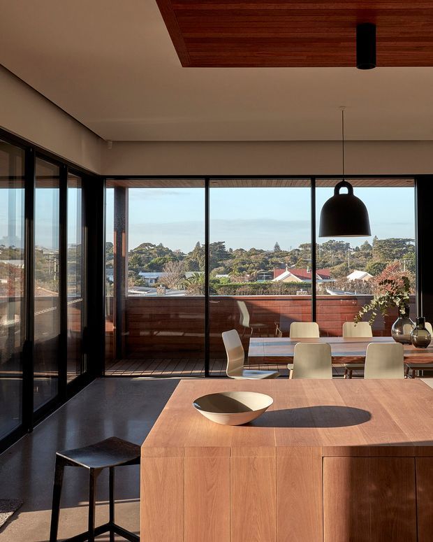 A warm contemporary dining and kitchen interior with expansive black-framed glazing overlooking a suburban landscape. The foreground features a large timber kitchen island or peninsula with a smooth wood finish, while a dining table and light upholstered chairs sit beside the windows. A dark stone or polished concrete floor, timber ceiling inset, black pendant lights, and dark-framed sliding or folding doors create a refined indoor-outdoor connection.