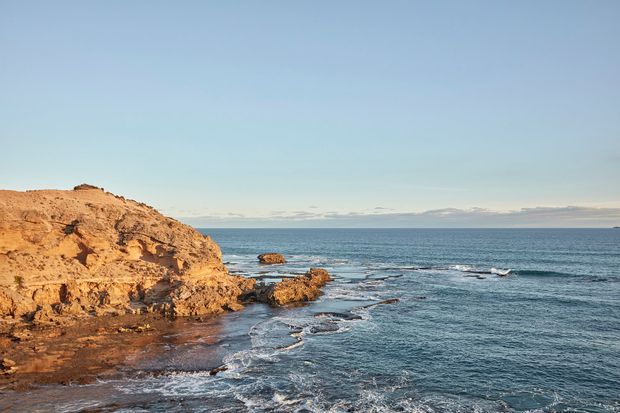 A wide coastal landscape view showing rocky ochre cliffs and a rocky shoreline meeting the blue ocean, with a calm horizon under a clear pale-blue sky. No building or designed architectural space is visible.