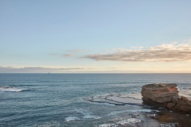 A wide coastal seascape viewed from the shoreline, showing calm blue-grey ocean stretching to the horizon, a rocky layered headland and low rock platforms in the foreground, and a pale sky with soft clouds. No building or interior space is visible.