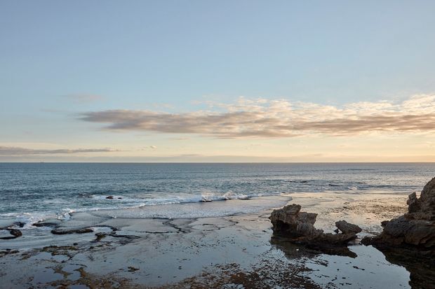 A tranquil coastal seascape viewed from a rocky shoreline, with calm ocean water stretching to the horizon, shallow tidal pools and dark rock formations in the foreground, and a softly lit blue and peach evening sky. No building or interior space is visible.