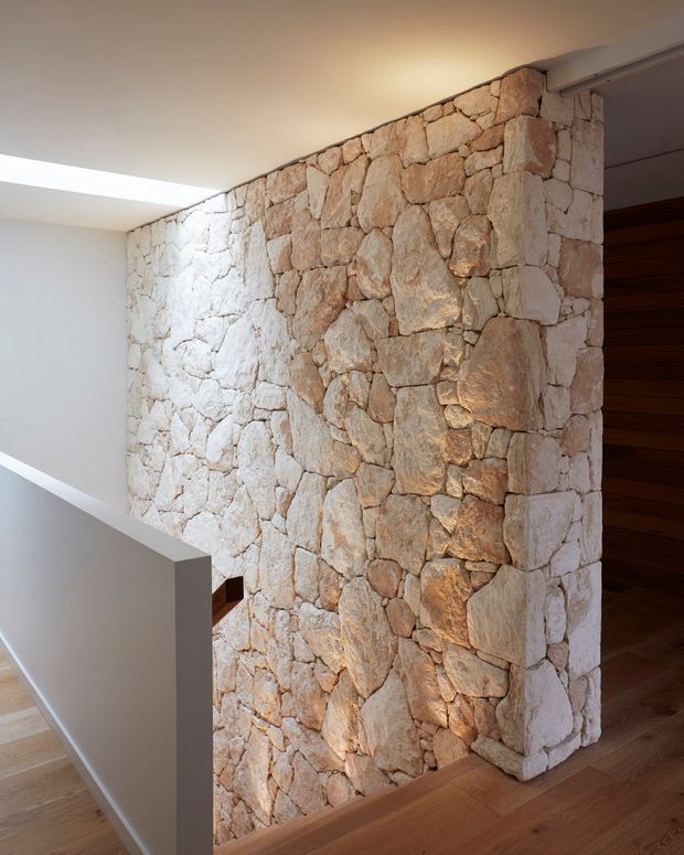 Interior architectural detail showing a rustic natural-stone feature wall built from irregular pale beige stones, forming a stairway or landing area. Warm timber flooring and a dark timber-clad doorway contrast with the white plaster ceiling and minimalist white stair balustrade. Recessed ceiling lighting highlights the textured masonry.
