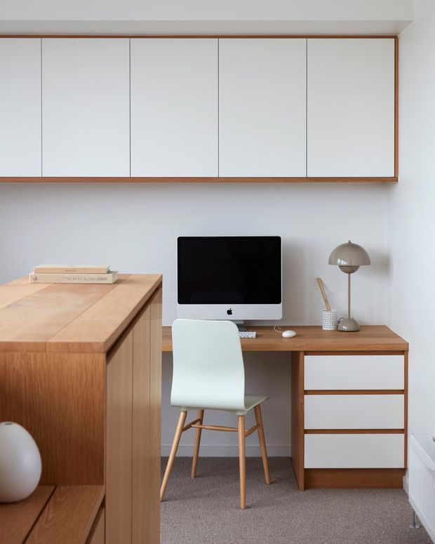 A compact, neatly arranged home office with a warm timber desk and integrated cabinetry. A white iMac sits on the desk beside a rounded metallic desk lamp and stationery, with a pale molded chair on wooden legs. Wall-mounted white storage cabinets framed in timber span the upper wall, while a large timber cabinet or shelving unit occupies the left foreground. The room has white walls, carpet flooring, and a calm minimalist mid-century-inspired feel.