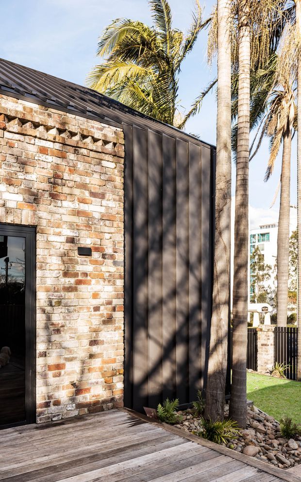 Exterior view of a contemporary building with a weathered exposed brick wall, tall dark metal-clad vertical panel, dark pitched metal roof, and black-framed doorway. A timber deck and landscaped garden with palm trees and river stones occupy the foreground, with neighboring urban buildings visible beyond.