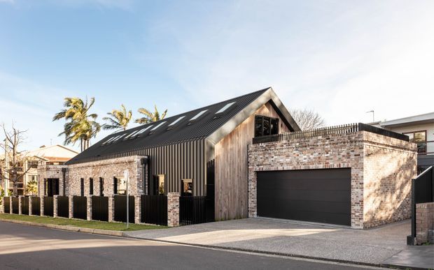 Street-facing exterior of a contemporary residential building with a steep gabled roof, dark standing-seam metal cladding, exposed timber-look gable wall, brick masonry sections, and an integrated double garage. Vertical black fencing and a paved driveway define the frontage, with neighbouring homes and palm trees in the background.