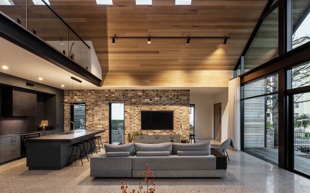Open-plan contemporary living area with a large grey sectional sofa facing a television mounted on an exposed brick feature wall. A dark kitchen island and cabinetry sit to the left, while full-height black-framed glazing opens to the exterior on the right. The room has a warm timber-lined ceiling with skylights, track lighting, polished concrete or terrazzo flooring, and a partial upper-level balcony or mezzanine.