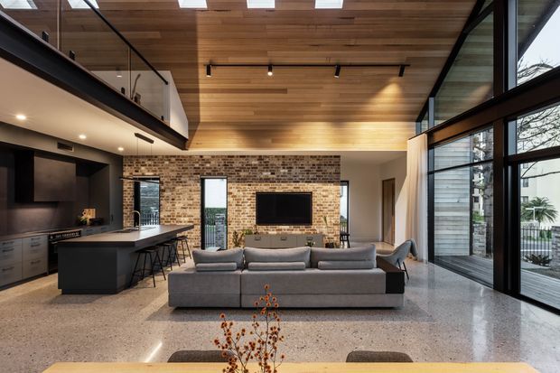 Open-plan contemporary living space combining a kitchen, dining area and lounge beneath a warm timber-lined ceiling. The room features a large grey sectional sofa, dark kitchen cabinetry with island seating, a brick feature wall with wall-mounted television, polished terrazzo flooring, extensive black-framed glazing, and a mezzanine level with a glass balustrade. Skylights and full-height windows provide abundant natural light.