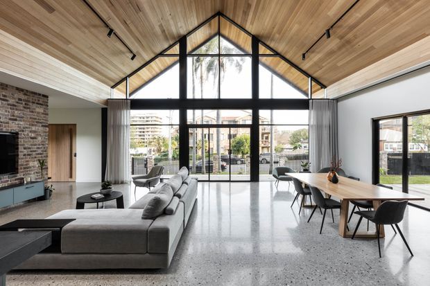 A bright contemporary open-plan living and dining space with a dramatic pitched timber-lined ceiling and full-height black-framed glazing. The room features polished concrete or terrazzo-style flooring, an exposed brick feature wall, a large grey sectional sofa, timber dining table, and minimalist black track lighting. The glazed gable end opens the room visually to the outdoors and provides abundant natural light.