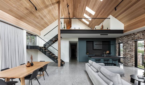 Open-plan contemporary living, dining and kitchen space with a dramatic timber-lined pitched ceiling, exposed brick wall, polished concrete flooring, and a mezzanine level with glass balustrading. The room includes a timber dining table, sectional sofa, dark kitchen cabinetry and a sculptural open stair.