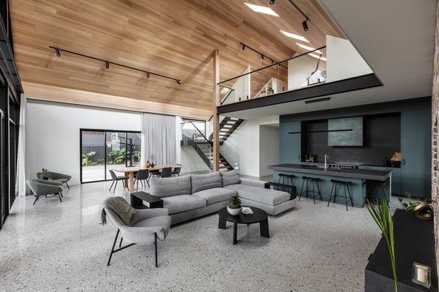A spacious contemporary open-plan living room with a double-height volume, polished terrazzo floor, timber-clad pitched ceiling, exposed timber posts, and a mezzanine level with black-framed glass balustrading. The room includes a large grey sectional sofa, lounge chairs, round coffee table, dining area, and a dark built-in media wall with a slim counter and stools. Full-height glazing opens toward an outdoor courtyard, while a stair connects to the mezzanine.