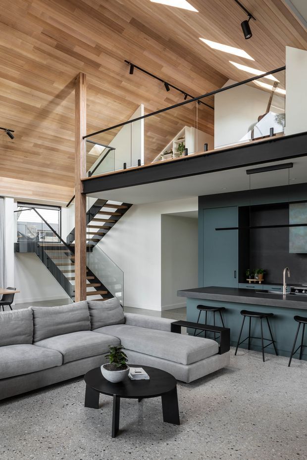 Contemporary open-plan interior featuring a living room and kitchen beneath a mezzanine. The space has a dramatic timber-lined sloped ceiling, exposed timber post, black metal stair and mezzanine balustrades, white walls, dark teal cabinetry, a grey sectional sofa, and a polished terrazzo floor. Skylights and generous glazing provide abundant natural light.