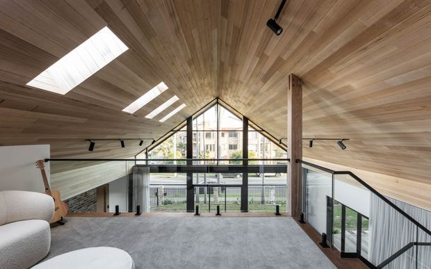 A contemporary loft-like interior with a dramatic pitched, timber-lined ceiling and multiple skylights. The upper-level space has a grey carpeted floor, glass balustrades overlooking the lower level, exposed timber supports, black-framed floor-to-ceiling glazing, and track lighting. A guitar and upholstered seating are visible, with an urban residential setting beyond the windows.