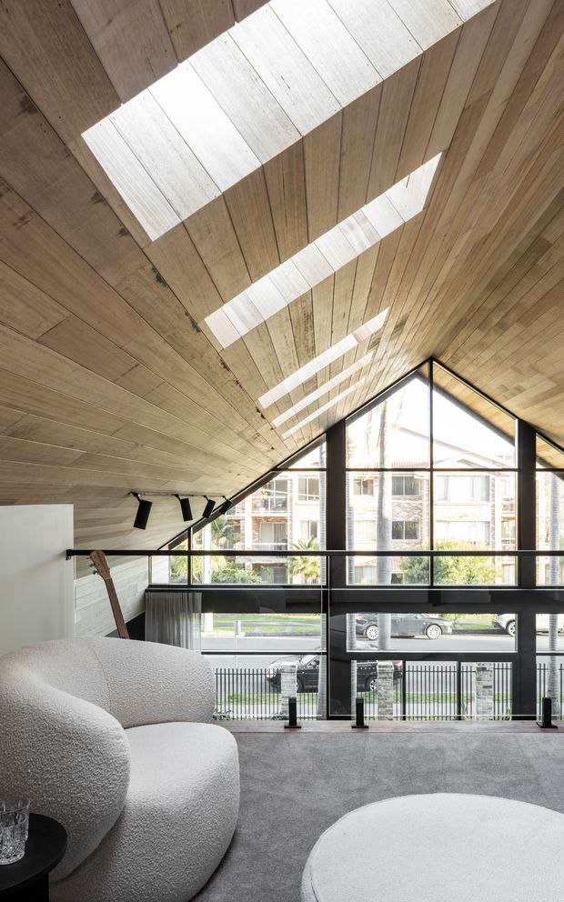 Contemporary double-height living room with a dramatic pitched timber-lined ceiling, large black-framed triangular glazing, and skylight-like roof openings that cast strong linear daylight across the wood. The room features a sculptural white boucle sofa, round white coffee table, grey carpet, black accents, and views to the street and neighbouring buildings.