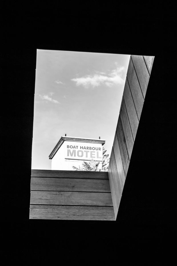 A high-contrast black-and-white architectural photograph looking upward through a narrow timber-lined opening or courtyard. The composition frames a clearly visible “Boat Harbour Motel” sign against the sky, with dark structural surfaces creating a dramatic geometric border.
