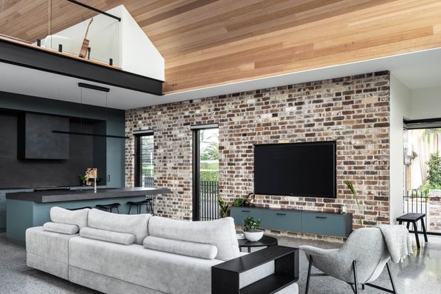 Open-plan contemporary living room with a large light-grey sectional sofa facing a wall-mounted television. The room features a prominent exposed brick feature wall, dark built-in cabinetry and entertainment joinery, a black kitchen island, extensive timber-lined ceiling, and high glazing opening toward an outdoor area. A glass balustrade and upper-level void are visible at left.