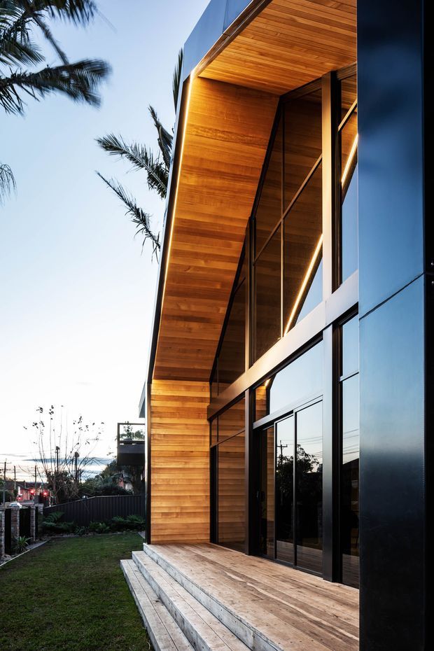 Exterior architectural photograph of a contemporary multi-level building with a dramatic projecting timber-lined soffit and matching timber wall cladding. Full-height black-framed glazing and dark metal facade panels create a strong vertical composition. A timber deck and steps lead along the facade, with palm trees and a residential streetscape in the background.