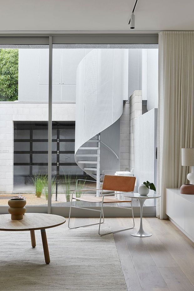 A bright, refined living room with floor-to-ceiling glazing overlooking a sculptural white spiral staircase and courtyard. The interior features pale timber flooring, a large neutral textured rug, a low round timber coffee table, a leather lounge chair, a small white side table, sculptural ceramics, minimal white built-in cabinetry, and full-height ribbed curtains. The space has a restrained contemporary aesthetic with strong geometric and curved architectural elements.