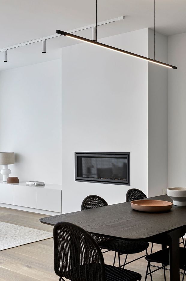 A minimalist open-plan living and dining area with a white built-in fireplace wall, low white media cabinetry, pale timber flooring, and a dark timber dining table surrounded by black woven chairs. Linear black pendant lighting and track spotlights add contemporary architectural detail, while neutral ceramics and a floor lamp provide restrained accents.