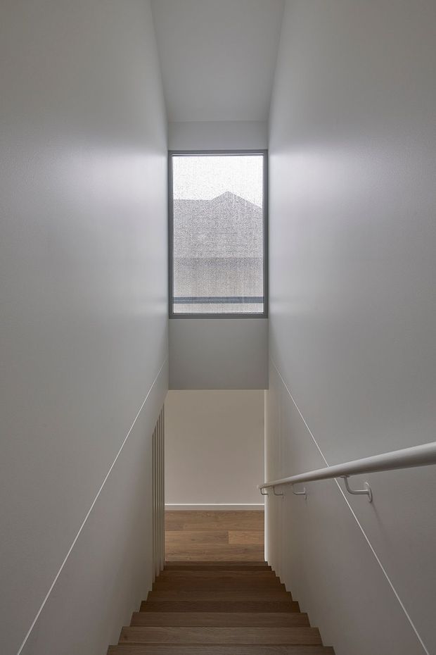 A narrow, minimalist stairway viewed from the upper landing, descending toward a wood-floored lower level. White walls and ceiling create a clean, enclosed vertical space, with a simple metal handrail on the right and a tall frosted or screened window at the far end providing soft natural light.