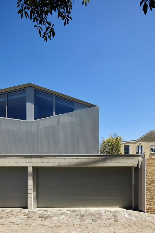 Street-facing architectural exterior of a contemporary multi-level residence, featuring a large double garage with dark sectional doors, pale masonry block construction, a projecting upper volume enclosed by blue-tinted glazing and translucent corrugated screening, and a paved stone driveway. A neighbouring traditional gabled house is visible in the background.