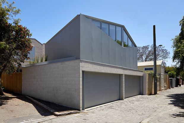 Street-level exterior view of a compact contemporary residential building, likely a townhouse or rear dwelling, with a two-car garage at ground level. The facade combines pale rendered or concrete-block walls with a sharply pitched, asymmetrical roof form and a large upper-level glazed window wall. A paved lane and neighbouring homes frame the urban residential setting.