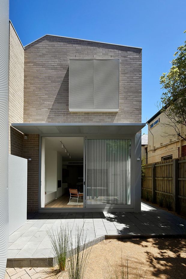 Rear exterior view of a compact two-storey urban dwelling or extension. The facade is formed from grey-brown brickwork with a simple shallow pitched roof, a large upper-level screened window, and a projecting flat-roofed ground-floor canopy enclosing full-height glazed doors. A paved courtyard and narrow landscaped strip occupy the foreground, with neighbouring buildings and fencing visible at the sides.
