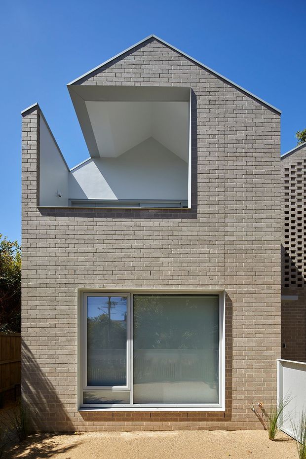 Front exterior view of a contemporary multi-level brick residence with a steep gabled roof and a dramatic recessed upper opening beneath the gable. The facade uses pale grey-brown brickwork, large frosted-glass windows with aluminium frames, and a perforated brick screen wall at the right. Strong sunlight and a clear blue sky emphasize the geometric form.