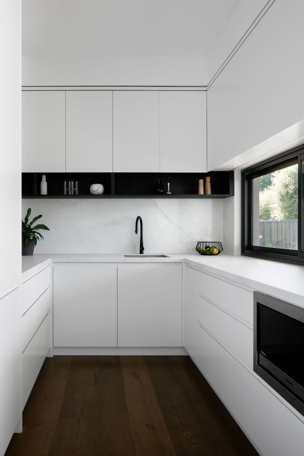 Compact contemporary white L-shaped kitchen with flat-panel cabinetry, a dark open display shelf, white veined stone backsplash and benchtops, black tapware and appliances, and dark timber flooring. A large black-framed window provides natural light and views to a fenced garden area.