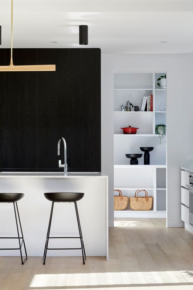 Bright, minimalist kitchen interior with a white island and slim black bar stools, a dramatic black timber-grain feature wall, integrated sink and curved chrome tap, white open shelving displaying cookware, books, plants and baskets, and pale timber flooring. A built-in oven is visible at right. Natural sunlight creates strong floor shadows.