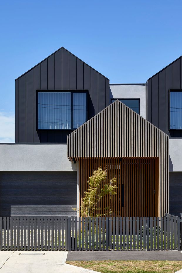 Front exterior view of a contemporary multi-level townhouse or residential building. The facade combines dark standing-seam metal cladding, pale rendered walls, large glazed windows, and a prominent timber vertical-slat entry canopy with a gabled form. A matching dark vertical-slat fence and driveway sit in the foreground under a clear blue sky.