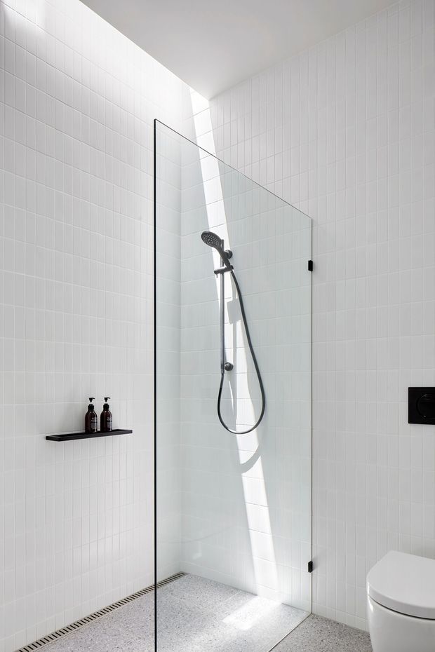 Bright minimalist walk-in shower bathroom with white vertically gridded wall tiles, a frameless glass shower screen, matte black shower fixtures and controls, a narrow black shelf with toiletries, a linear floor drain, and a pale terrazzo-style floor. Strong natural light enters from a high opening or skylight, creating a clean, airy atmosphere.