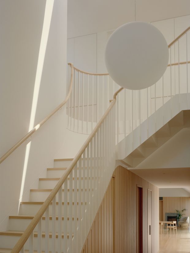 A bright, refined interior view of a sculptural staircase and landing. White-painted balustrades with slender vertical spindles and pale timber handrails frame the stairs, while warm vertical timber wall panelling and pale timber flooring add texture. A large spherical pendant light hangs in the stair void, and an open hallway with glimpses of dining or living furniture is visible beyond.