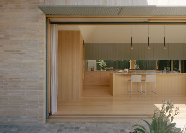 View from an exterior paved area into a contemporary open-plan kitchen through a fully opened glazed sliding door. The kitchen features extensive light timber cabinetry, a large island with seating for three, dark green vertical wall panelling, pendant lights, and pale timber flooring. The exterior facade is clad in light beige brick with a projecting ribbed metal awning.