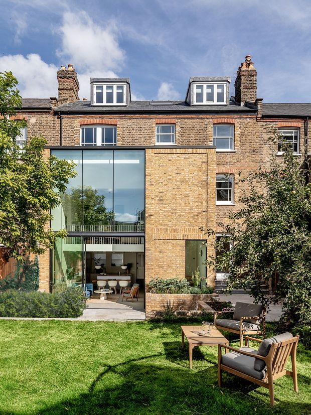 Rear exterior view of a multi-storey brick townhouse or terrace-style residence with a contemporary glazed extension opening onto a landscaped lawn and outdoor seating area. The original upper facade features dark brickwork, pitched roof sections, dormer windows, chimneys, and traditional sash-style windows, while the extension has large floor-to-ceiling glazing, a projecting upper-level balcony, and brick detailing. The scene is bright and leafy with a refined residential garden setting.