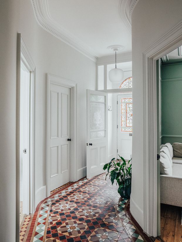 A bright, narrow residential hallway with ornate white plaster cornices, detailed panelled doors and trim, a patterned encaustic-style tiled floor, and a stained-glass entry door and fanlight. A potted plant sits along the curved corridor, with a glimpse of a green-accented living room through the doorway on the right.