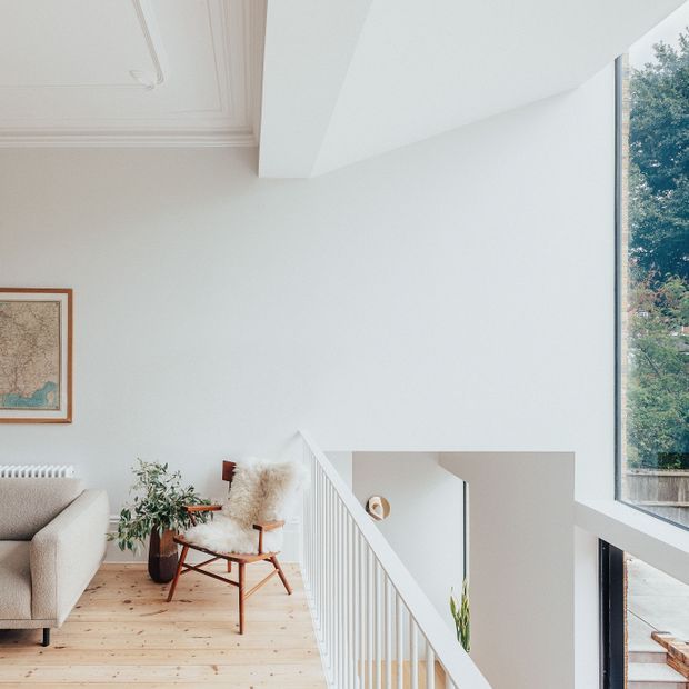 A bright, minimalist upper-level living area overlooking a stairwell. The space features white walls and ceiling detailing, pale timber floorboards, a cream sofa, a timber-framed lounge chair with a sheepskin throw, a potted plant, framed artwork, and a large vertical window with views to greenery. A white balustrade and open stair void define the foreground.
