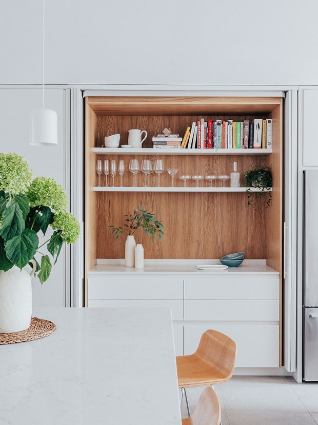 Bright contemporary kitchen featuring a large pale stone island in the foreground, integrated white cabinetry, and a recessed open display cabinet lined in warm timber veneer. The shelving holds books, glassware, crockery and decorative plants, with timber dining chairs and a stainless-steel refrigerator visible at right.