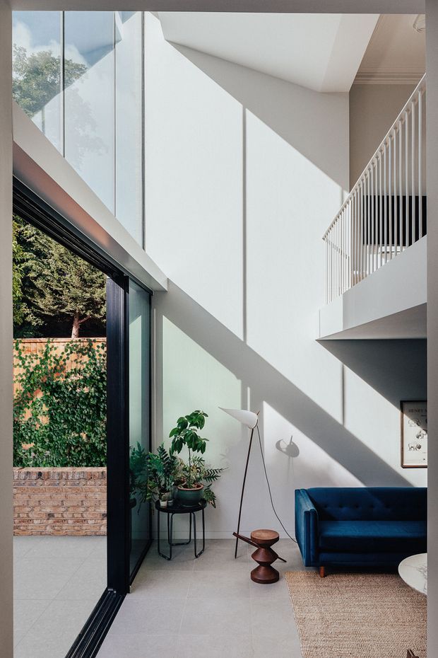 A bright, double-height contemporary living room with large floor-to-ceiling glazed doors opening to a leafy courtyard. Strong geometric sunlight and shadows fall across smooth white walls and pale flooring. The room includes a deep blue sofa, sculptural floor lamp, indoor plants, a small dark timber side table, and a woven rug. A white balustrade overlooks the space from an upper level, emphasizing the open vertical volume.