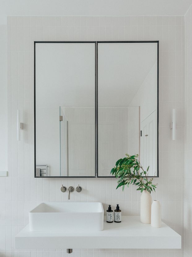 A bright, minimalist bathroom vanity with a white rectangular vessel basin set on a broad white benchtop. The wall is clad in narrow vertical white tiles, with two large black-framed mirrored cabinets above the basin. Wall-mounted brushed-metal tapware, neutral bathroom accessories, and a simple vase with green foliage complete the calm, monochromatic composition.