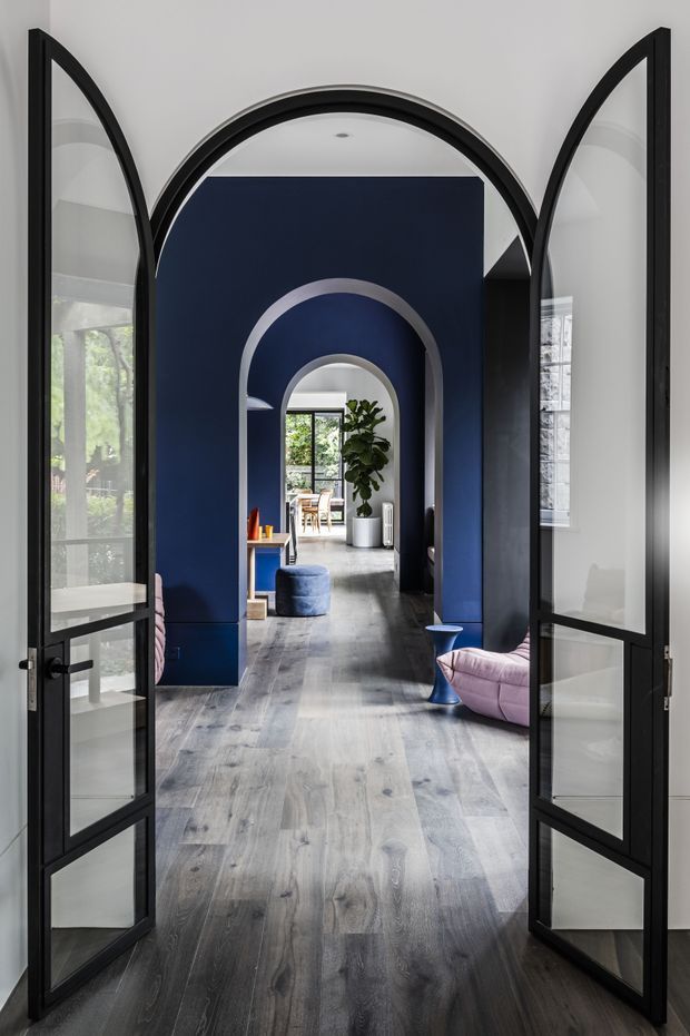 A dramatic interior view through open black-framed glazed double doors into a sequence of arched openings. Deep navy feature walls and arch reveals contrast with pale grey timber flooring, while curved pink lounge seating and blue accent stools add colour. Glazing at the sides and rear brings in greenery and daylight, creating a refined, bold transitional space.
