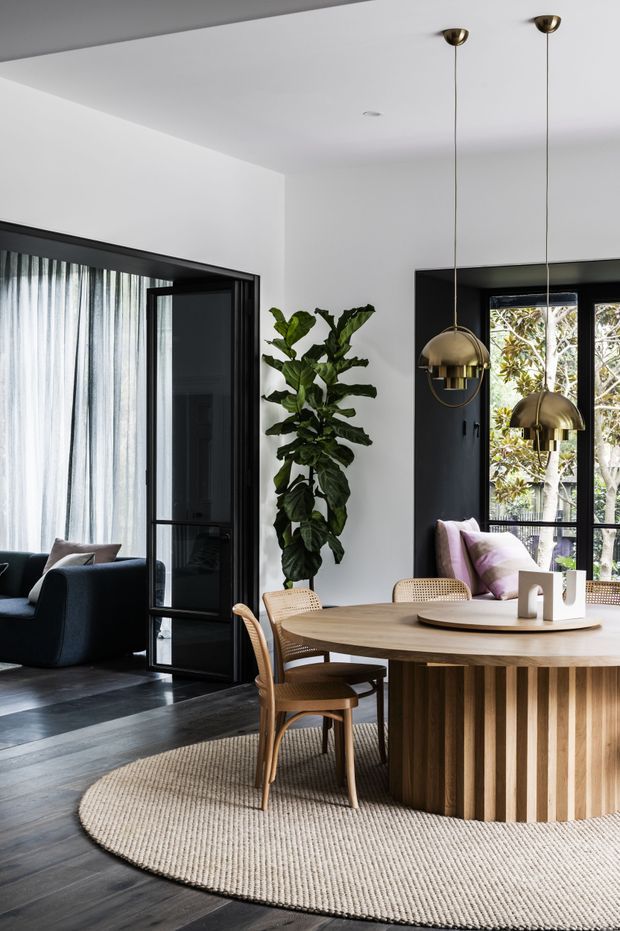 A refined dining area with a large round fluted timber dining table, woven dining chairs, and two sculptural brass pendant lights. The room has white walls and ceiling, dark timber flooring, black-framed glazed doors and windows, and a neutral woven circular rug. A black-framed opening connects to a darker living room with sheer curtains and a dark sofa. A tall indoor plant and soft cushions add natural and muted colour accents.