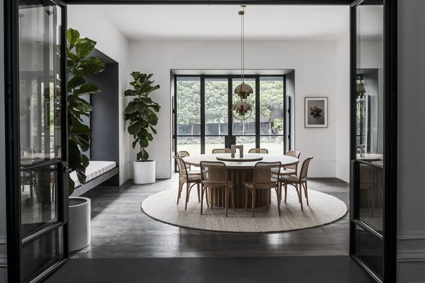 A refined dining room viewed through open black-framed glazed doors. The room features a round wood dining table with matching chairs, a large oval neutral rug, dark timber flooring, white walls, black-framed windows and doors, pendant lighting, built-in window seating, indoor plants, and a framed artwork. The space has a balanced, contemporary look with strong contrast between light walls and dark architectural elements.