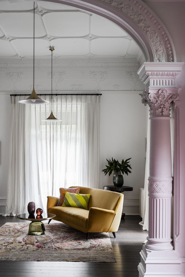 A highly detailed, ornate living room in a historic-style interior. The room features a decorative coffered ceiling, elaborate plaster cornices and floral mouldings, and a large pink-painted classical column with an ornate capital and base. Full-height sheer curtains frame a tall window, while a mustard-yellow velvet armchair, patterned area rug, low coffee table, pendant lights, and indoor plant create a refined eclectic sitting area. The palette is predominantly pale pink, white, mustard, and dark timber.