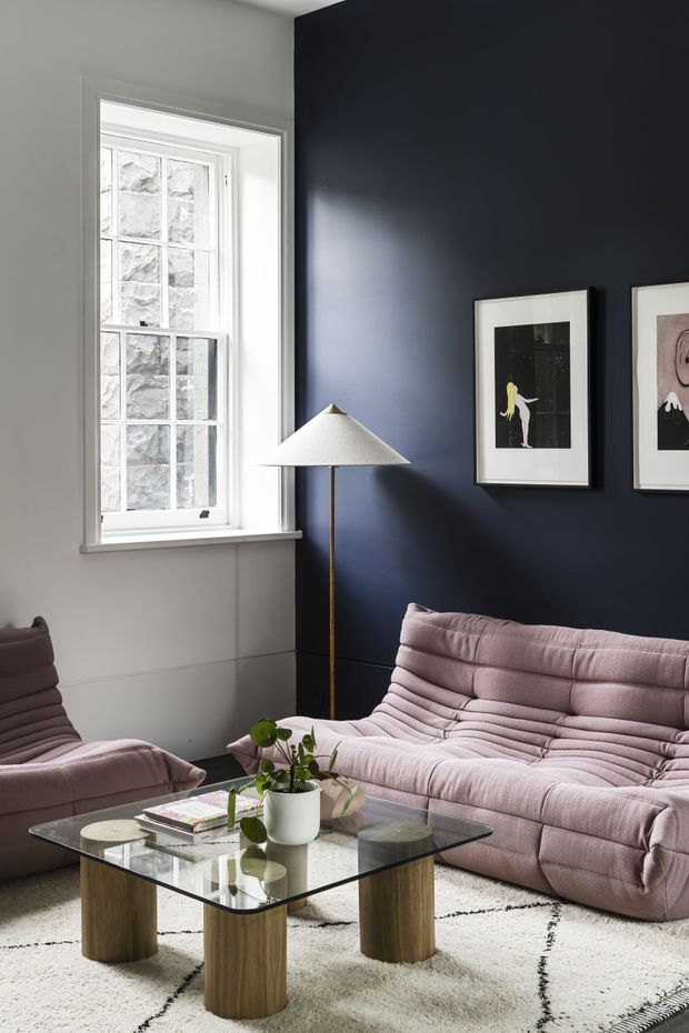 A refined living room with a deep navy feature wall, pale grey-white surrounding walls, and a tall multi-pane window. A sculptural floor lamp, framed artwork, and a blush-pink modular sofa and armchair surround a square glass coffee table with cylindrical timber legs. A light patterned rug and small potted plant complete the softly lit, contemporary setting.